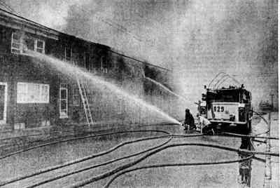 Firefighters directing hose streams onto a burning two-storey building, ladder raised, engine 129 at the kerb