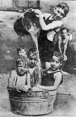 Children in bathing suits crowded into a galvanised washtub while an older boy pours water from a bucket