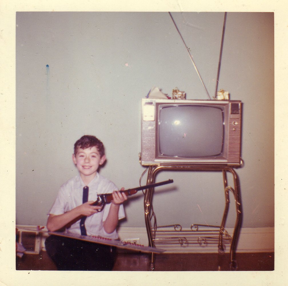 Michael Becoskie with Christmas air rifle in dining room. Remote controlled TV, rabbit ears antenna, no UHF antenna.