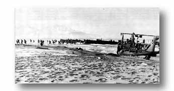 Infantrymen pour onto the beaches as bulldozer, later hit by enemy shell, works to clear beach obstacles