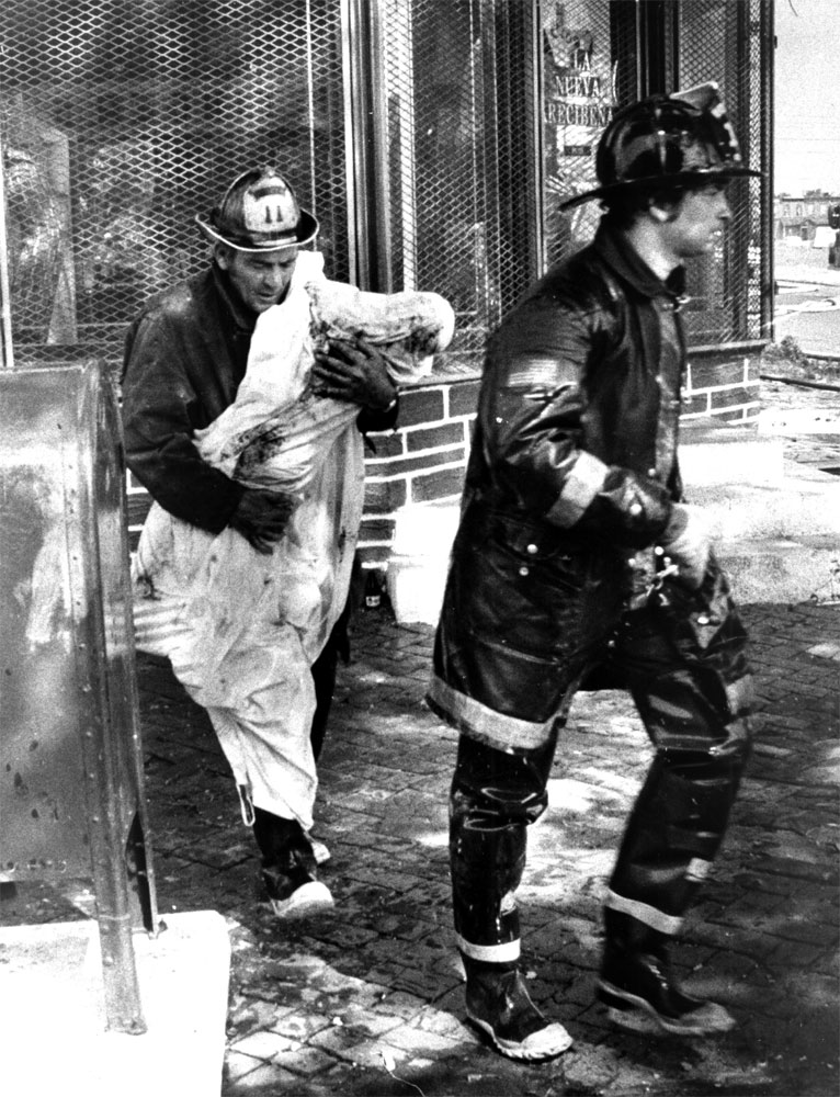 Captain Jim McGrory removes the body of a child killed when a house was fire-bombed at 600 Elm Street, July 4, 1974